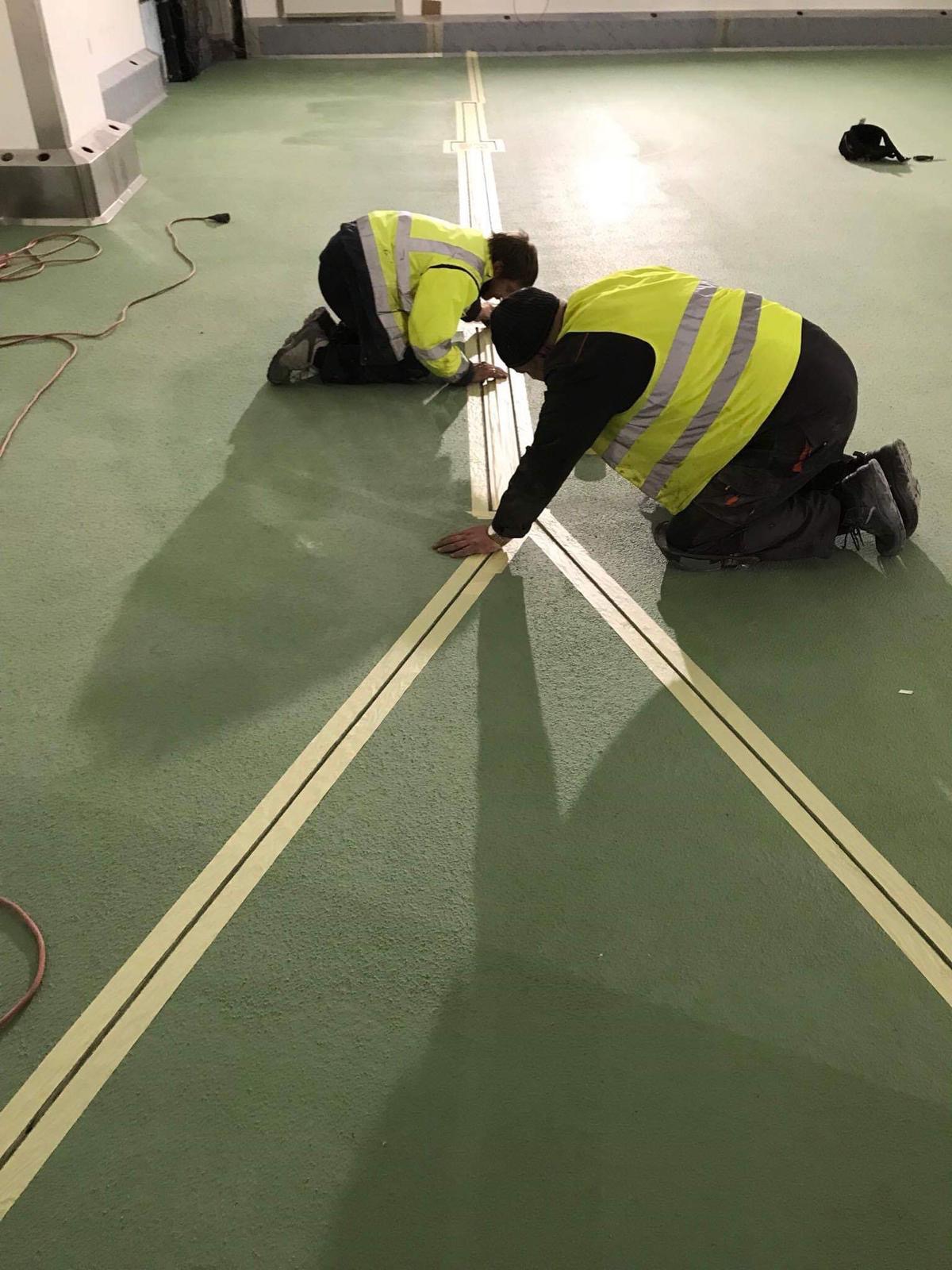 Dwóch pracowników w odblaskowych kamizelkach, na kolanach, aplikuje taśmę wyznaczającą linie na zielonej, szorstkiej posadzce przemysłowej w hali.
