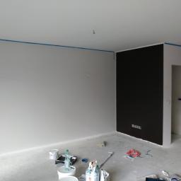 Capital Uslugi Budowlane - Pokój w trakcie malowania: szare ściany z niebieską taśmą malarską przy suficie, jedna ściana pomalowana na ciemny brąz, widoczne narzędzia i materiały malarskie na podłodze.