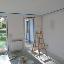 Capital Uslugi Budowlane - Wnętrze w trakcie prac wykończeniowych: ściany przygotowane do malowania, zabezpieczone taśmą malarską przy suficie i oknach, drabina malarska, widoczne ślady szpachlowania, okna balkonowe...
