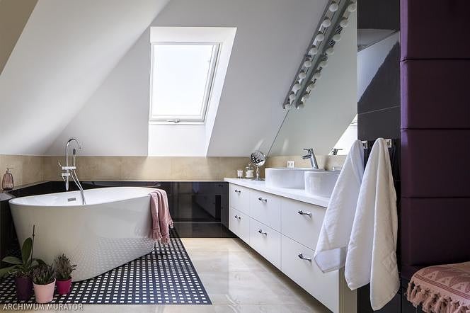 Nowoczesna łazienka na poddaszu z oknem dachowym, białą wanną wolnostojącą, podwójną umywalką nablatową i czarno-białą mozaiką na podłodze.