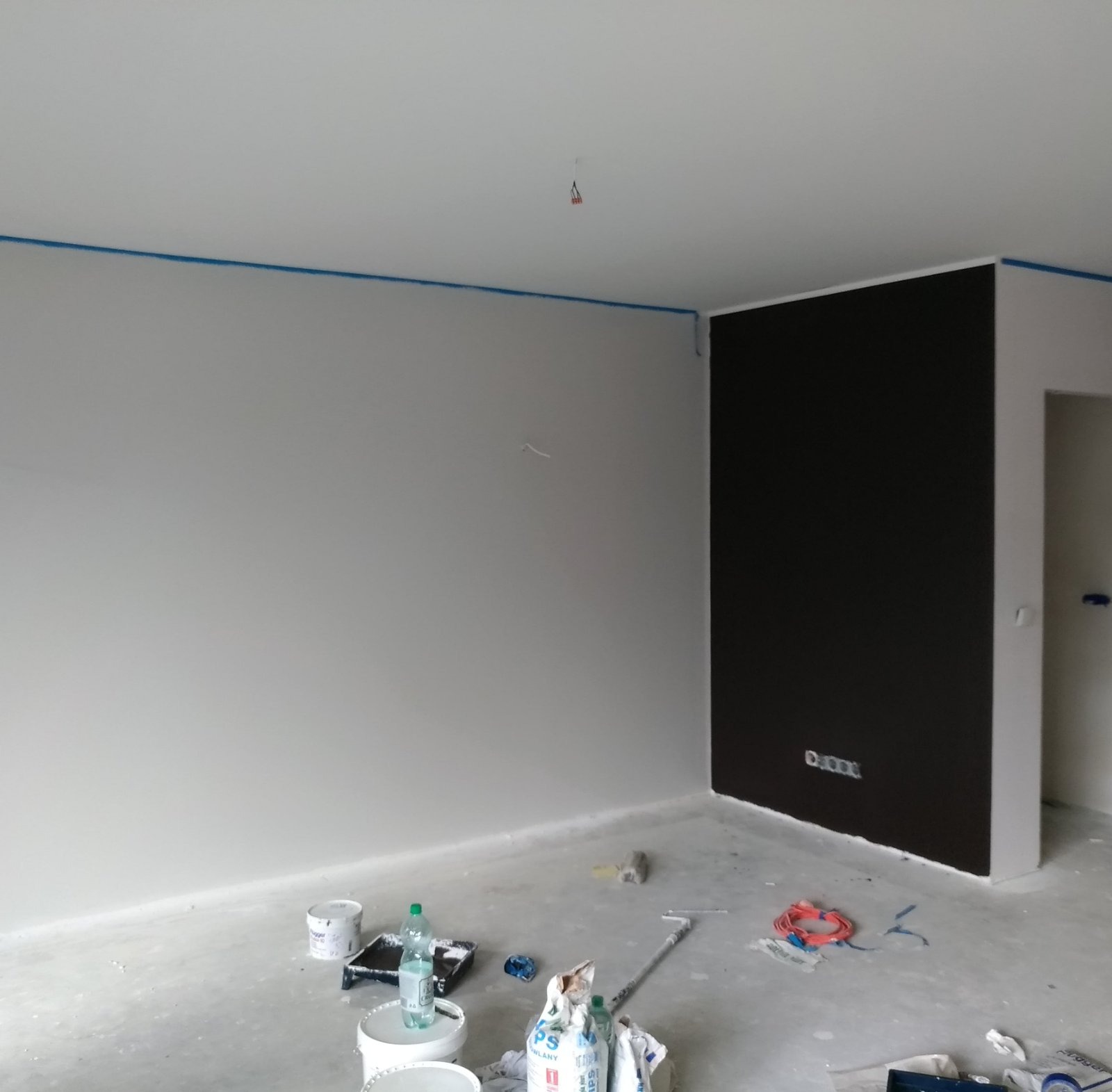 Pokój w trakcie malowania: szare ściany z niebieską taśmą malarską przy suficie, jedna ściana pomalowana na ciemny brąz, widoczne narzędzia i materiały malarskie na podłodze.