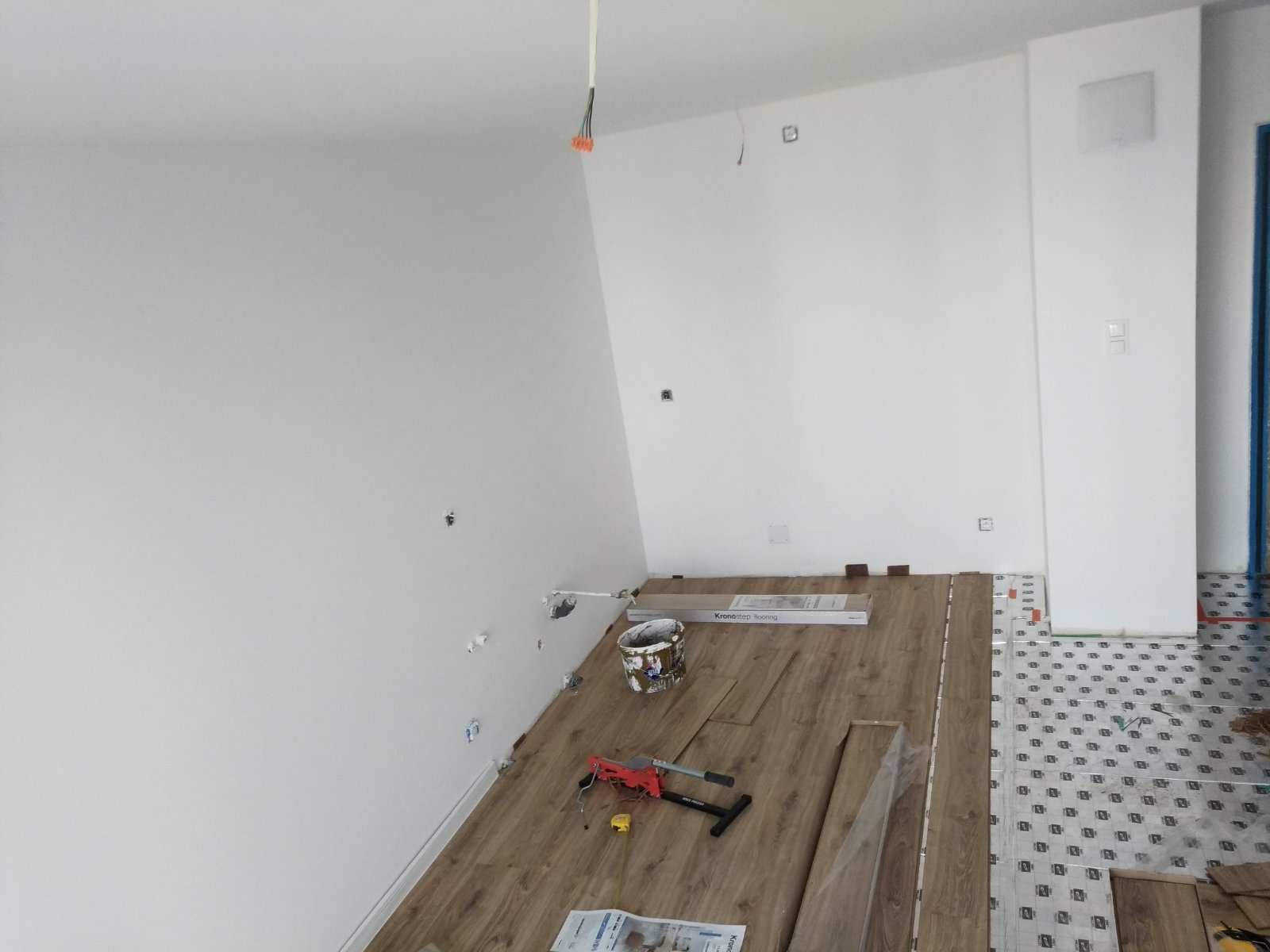 Montaż paneli podłogowych w pokoju, widoczne narzędzia, panele Kronostep, puszka po farbie i częściowo ułożona podłoga.