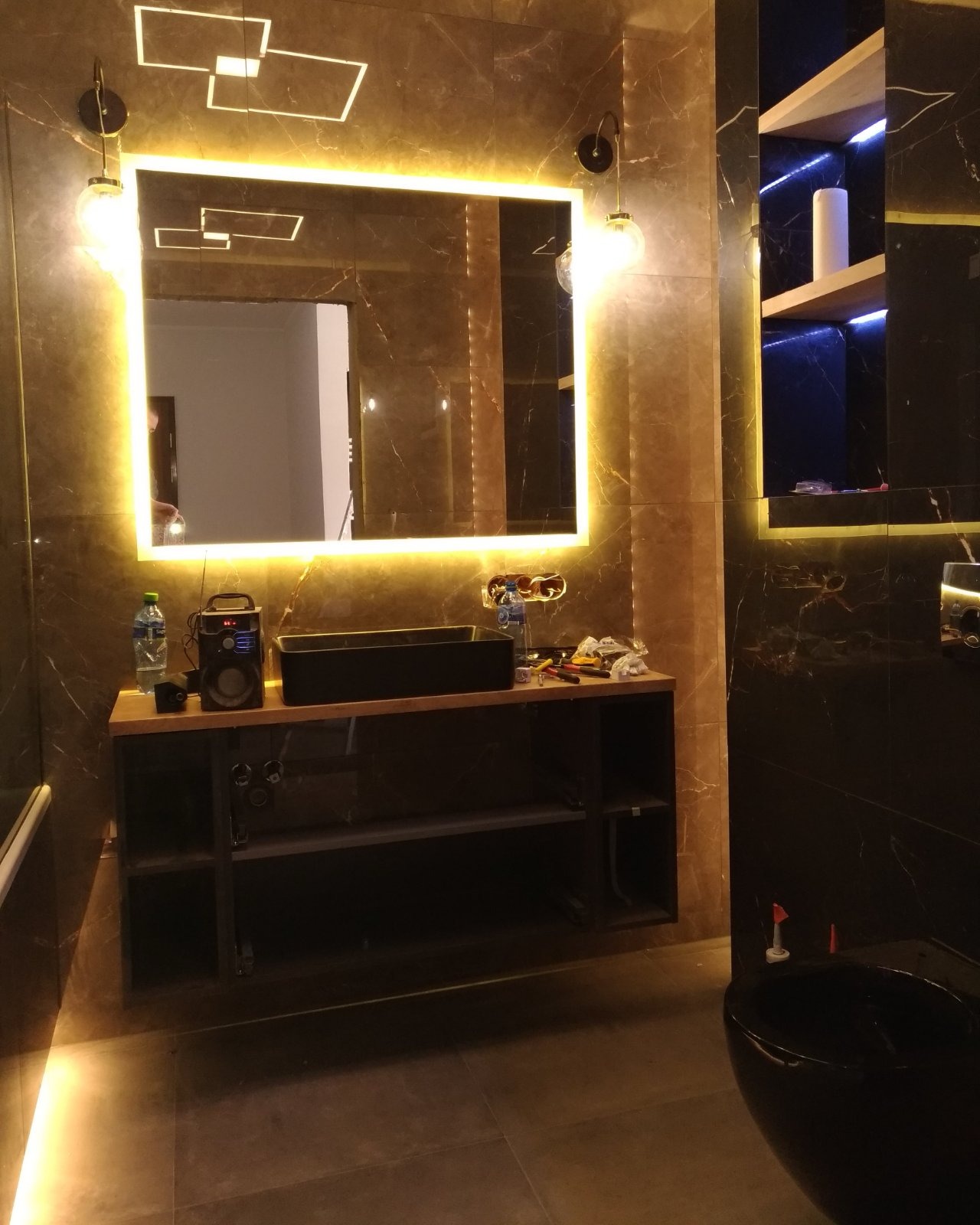 Nowoczesna łazienka z marmurowymi płytkami, podświetlanym lustrem, czarną umywalką nablatową i wiszącą toaletą. Na blacie obok umywalki stoją narzędzia i akcesoria, sugerujące trwające prace...