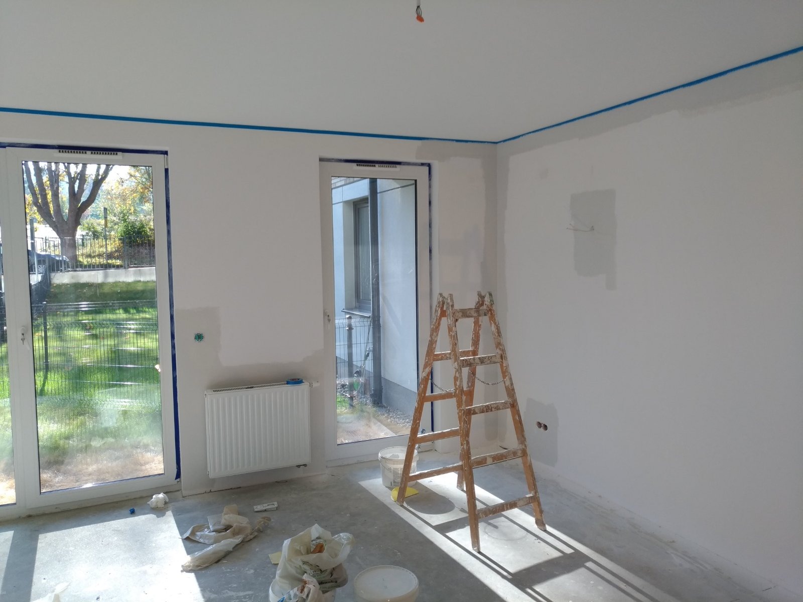 Wnętrze w trakcie prac wykończeniowych: ściany przygotowane do malowania, zabezpieczone taśmą malarską przy suficie i oknach, drabina malarska, widoczne ślady szpachlowania, okna balkonowe...
