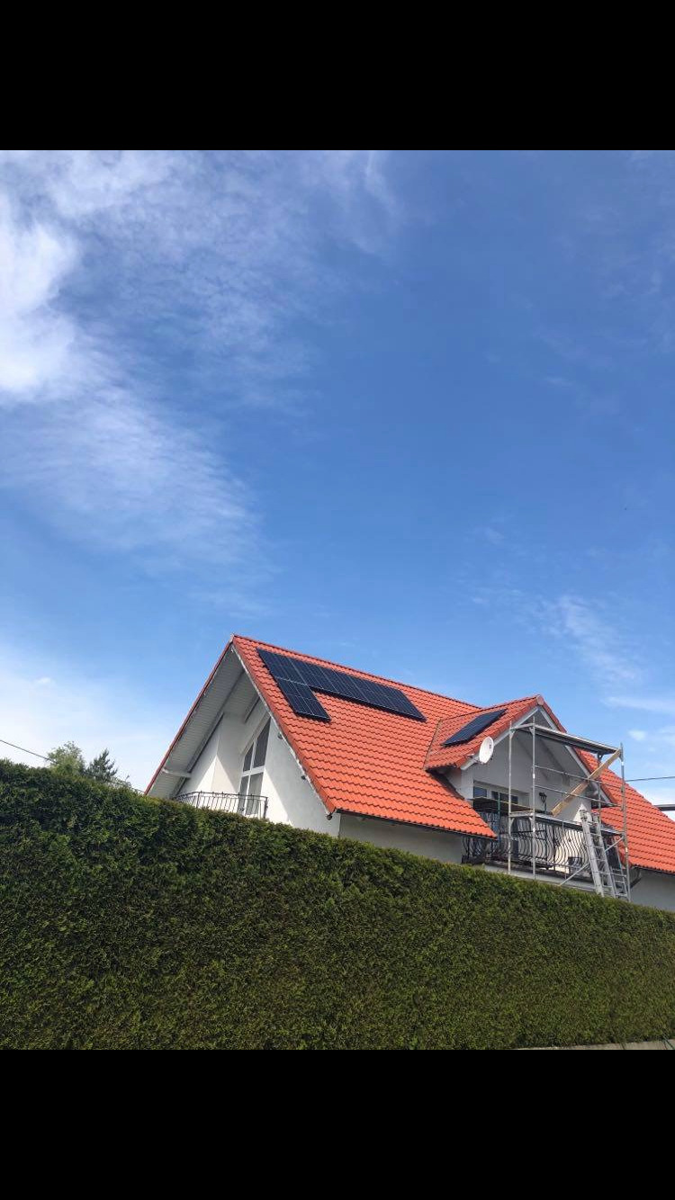 Dom jednorodzinny z pomarańczową dachówką i zainstalowanymi panelami słonecznymi, widoczne rusztowanie przy balkonie, ujęcie zza wysokiego żywopłotu, błękitne niebo.