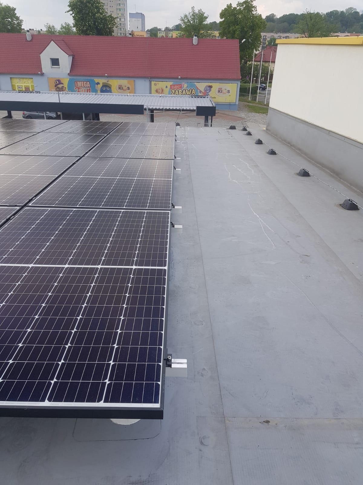 Panele słoneczne zamontowane na płaskim dachu budynku z widocznymi elementami mocującymi i oddalonym budynkiem w tle.