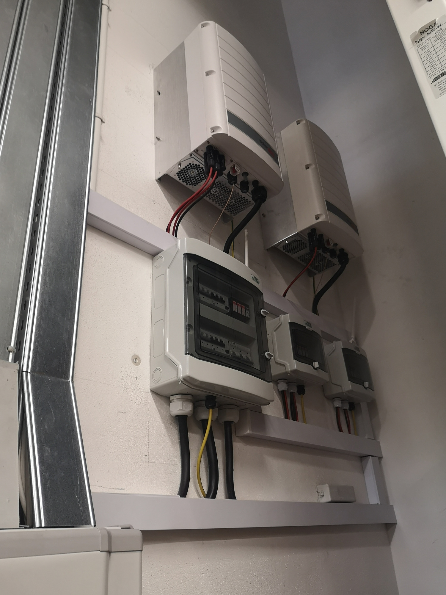 Dwa inwertery fotowoltaiczne zamontowane na ścianie, poniżej skrzynka rozdzielcza z przewodami w kolorach czarnym, czerwonym i żółtym, widoczne również metalowe koryta kablowe.