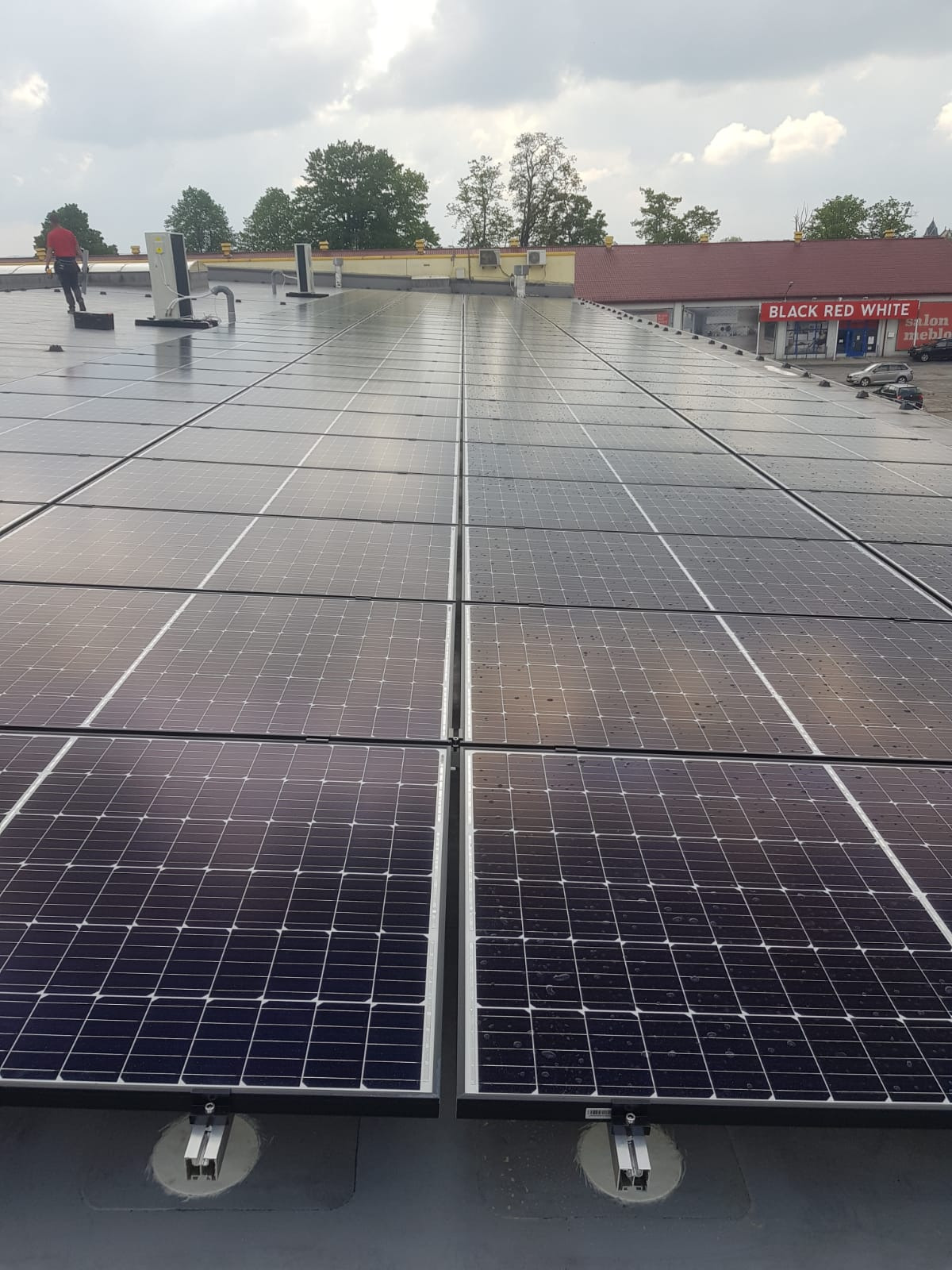 Rozległa instalacja paneli słonecznych na dachu budynku komercyjnego, widoczne mocowania, krople wody na panelach, w tle budynek z szyldem 'Black Red White' oraz sylwetka osoby w czerwonej koszulce.