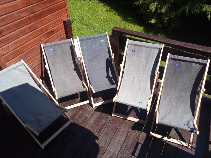 Pięć drewnianych leżaków z szarym płótnem, ustawionych na drewnianym tarasie w słoneczny dzień, widoczne z góry, z częściowym cieniem rzucanym przez jeden z leżaków.