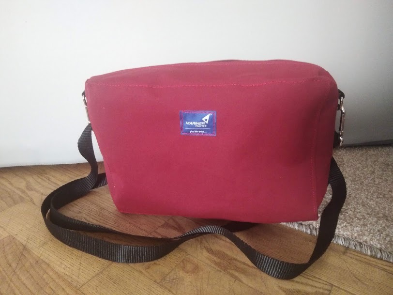 Burgundowa torba z materiału z czarnym paskiem na ramię, leżąca na drewnianej podłodze, z widocznym logo 'Mariner Nights'.