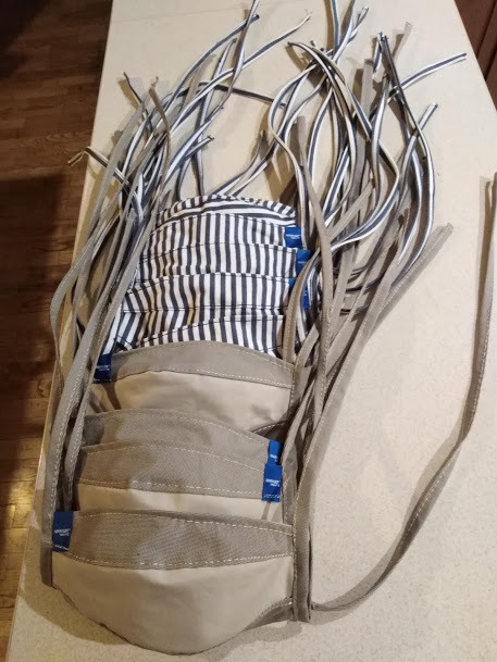 Stos maseczek ochronnych: trzy beżowe z szarym paskiem i jedna w czarno-białe paski, wszystkie z niebieskimi metkami, z wystającymi sznurkami.