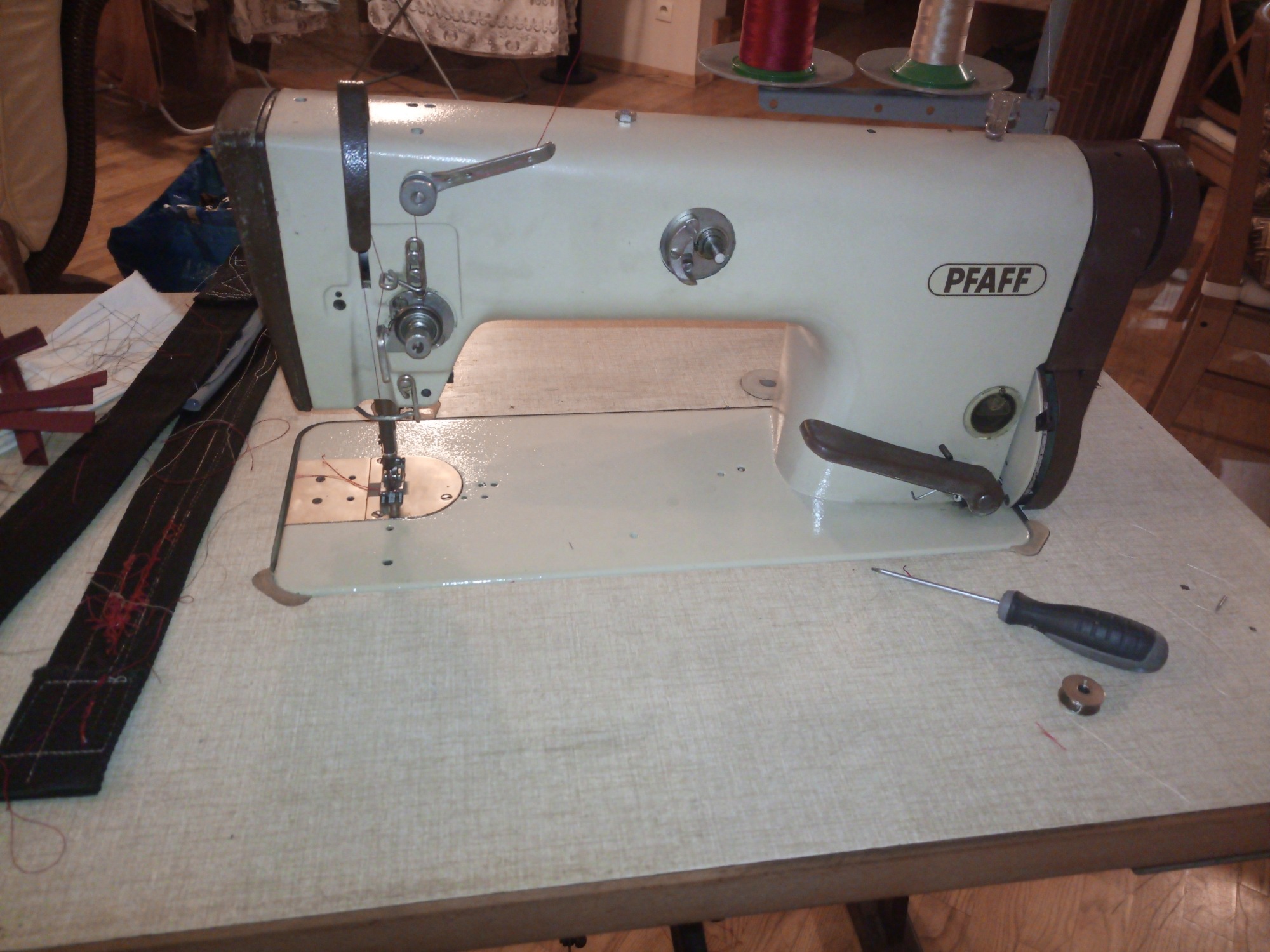 Vintage maszyna do szycia marki Pfaff na stole z fragmentem szytego paska, śrubokrętem i szpulką.