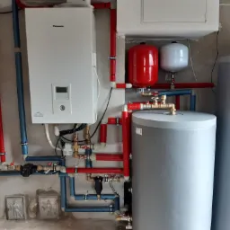 Biała pompa ciepła Panasonic Aquarea z licznymi rurami (czerwone, niebieskie) i zbiornikiem akumulacyjnym w kotłowni. Na ścianie widoczne ozdobne, metalowe płaskorzeźby.