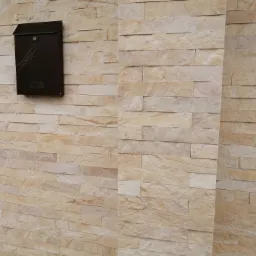 Elewacja z piaskowca w odcieniach beżu i kremu ułożona w poziome pasy, z zamontowaną nowoczesną, brązową skrzynką na listy z delikatnym wzorem.