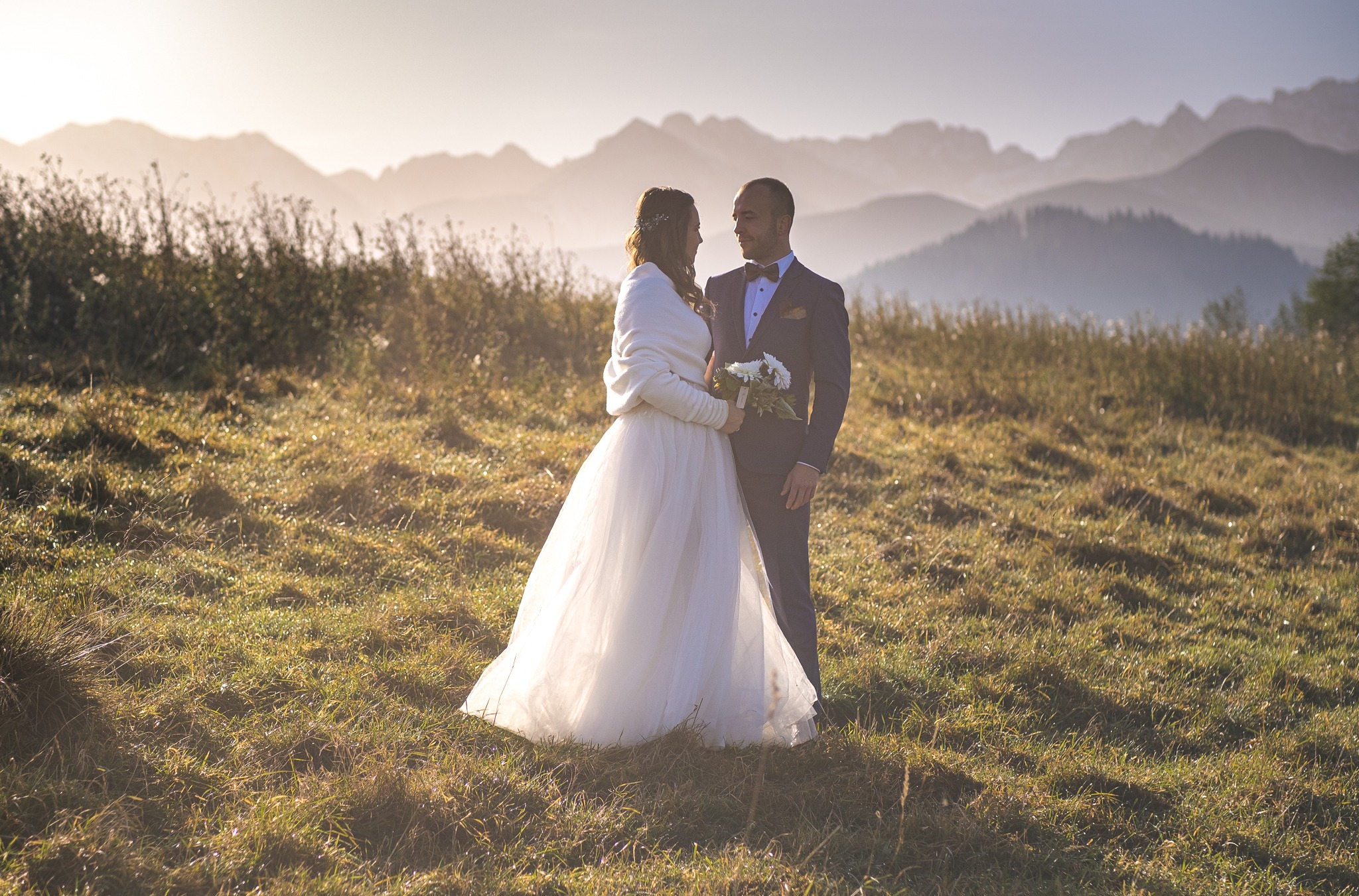 Para młoda w plenerze, panna młoda w białej sukni i kremowym okryciu, pan młody w granatowym garniturze z muszką, w tle góry w lekkiej mgle, złote światło zachodzącego słońca.