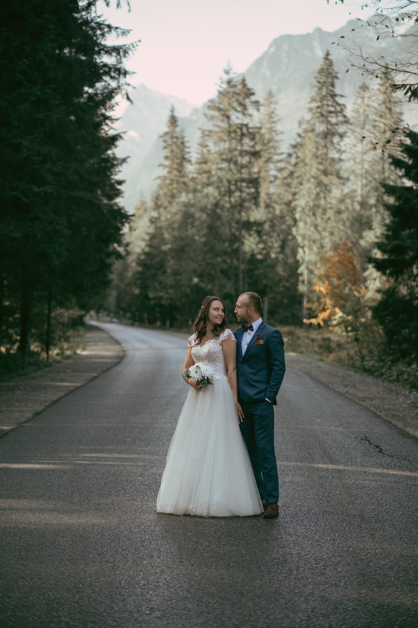 Para młoda w strojach ślubnych stoi na asfalcie drogi otoczonej lasem iglastym z górami w tle, panna młoda trzyma bukiet białych kwiatów.