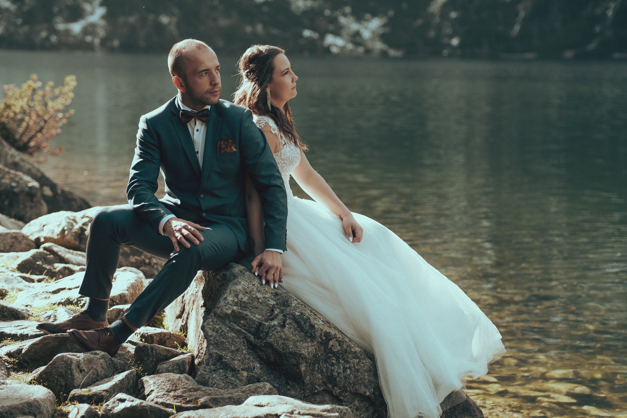 Elegancka para młoda siedzi na skale nad brzegiem jeziora, on w ciemnozielonym garniturze z muszką, ona w białej sukni ślubnej z koronkową górą, trzymają się za ręce, oboje patrzą w dal.