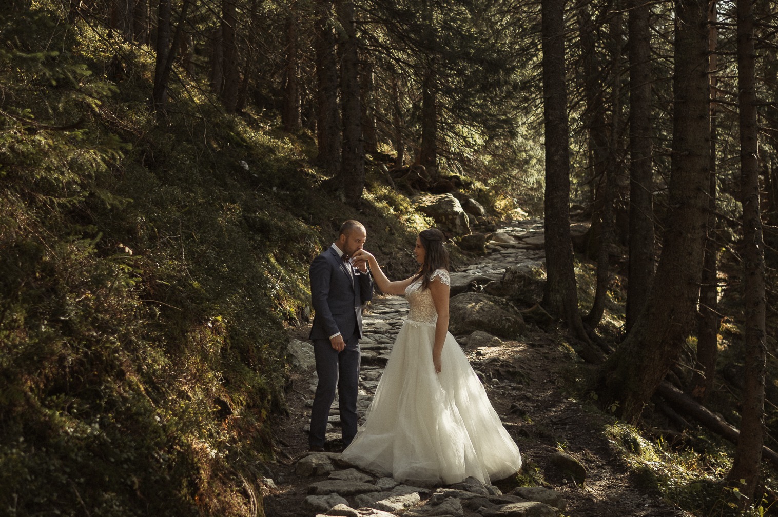 Para młoda w lesie na kamienistej ścieżce: pan młody w granatowym garniturze całuje dłoń panny młodej w białej sukni z koronkową górą, wysokie drzewa i leśna roślinność w tle.