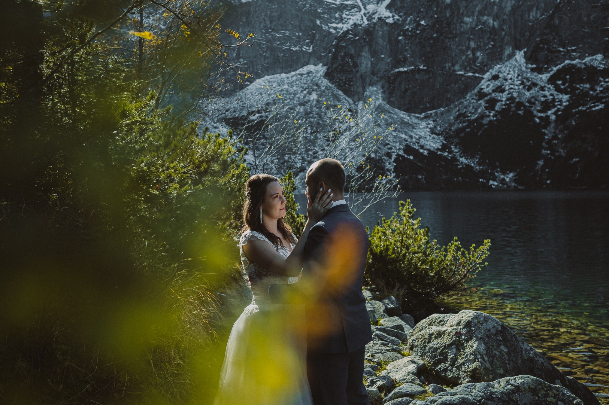 Para młoda w malowniczej scenerii górskiego jeziora, panna młoda w białej sukni z koronkową górą, pan młody w ciemnym garniturze, delikatny dotyk dłoni na twarzy, kamienisty brzeg i zielona...