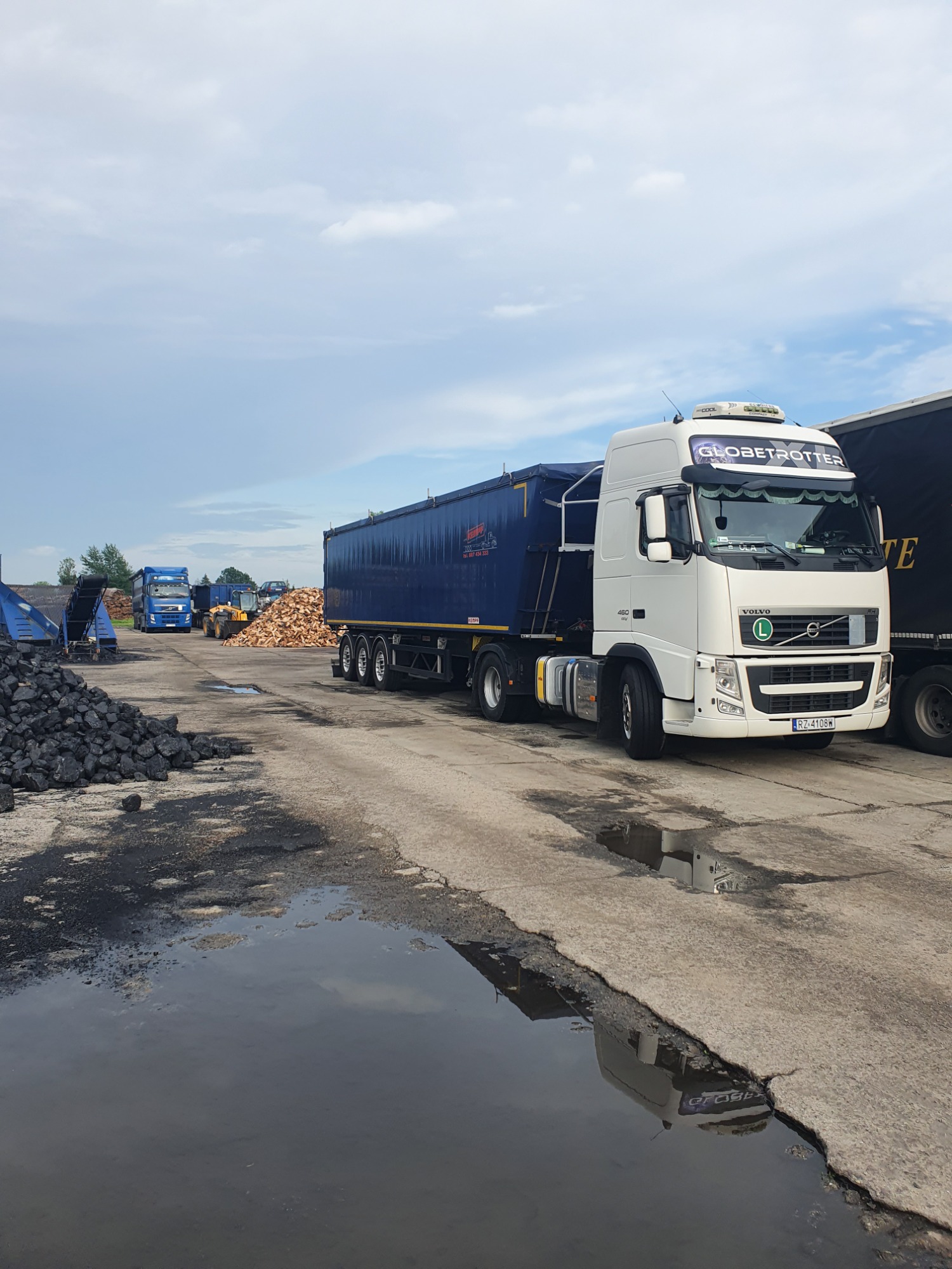 Ciężarówka Volvo Globetrotter z niebieską naczepą na placu składowym, w tle hałda węgla i stosy drewna, widoczne odbicie w kałuży.