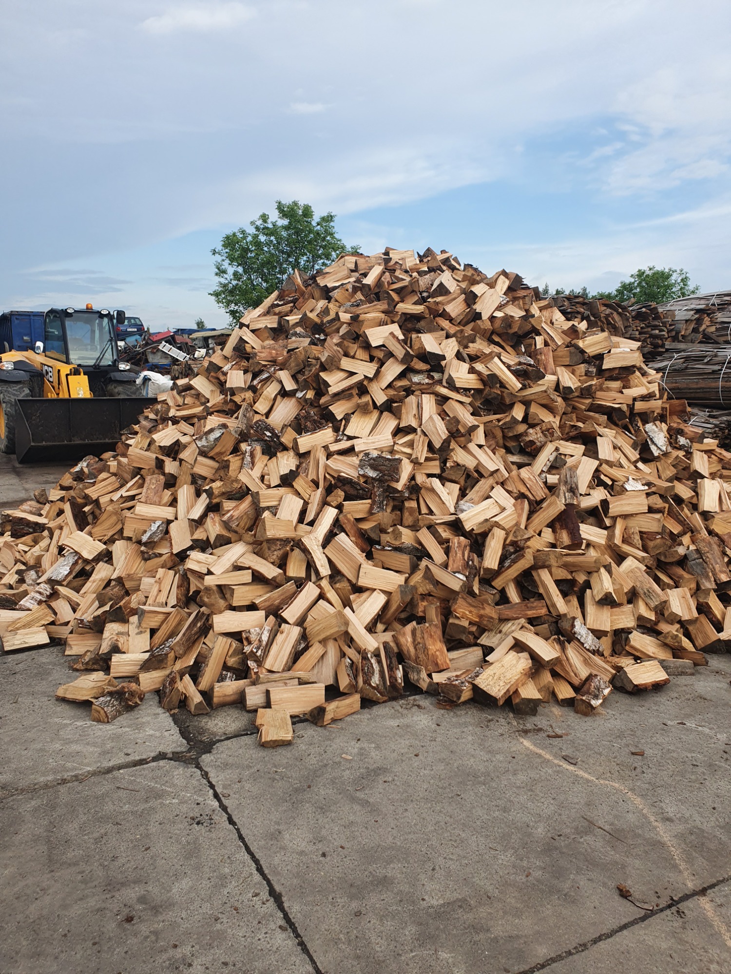 Stos pociętego drewna na betonowym placu, w tle widoczny traktor i drzewa pod błękitnym niebem.