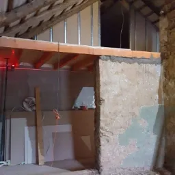 Adaptacja poddasza: widoczne elementy konstrukcyjne, drewniane belki stropowe, ściany z surowego tynku, metalowe profile oraz płyty gipsowo-kartonowe w trakcie montażu, poziomica laserowa...