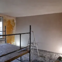 Pokój w trakcie remontu z rusztowaniem, drabiną i wiadrami z farbą, ściana częściowo pokryta nową warstwą, druga ściana z odsłoniętą starą warstwą farby.