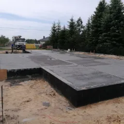 Wylany betonowy fundament pod budynek z widocznym szalunkiem i pracownikami przy betoniarce w tle.