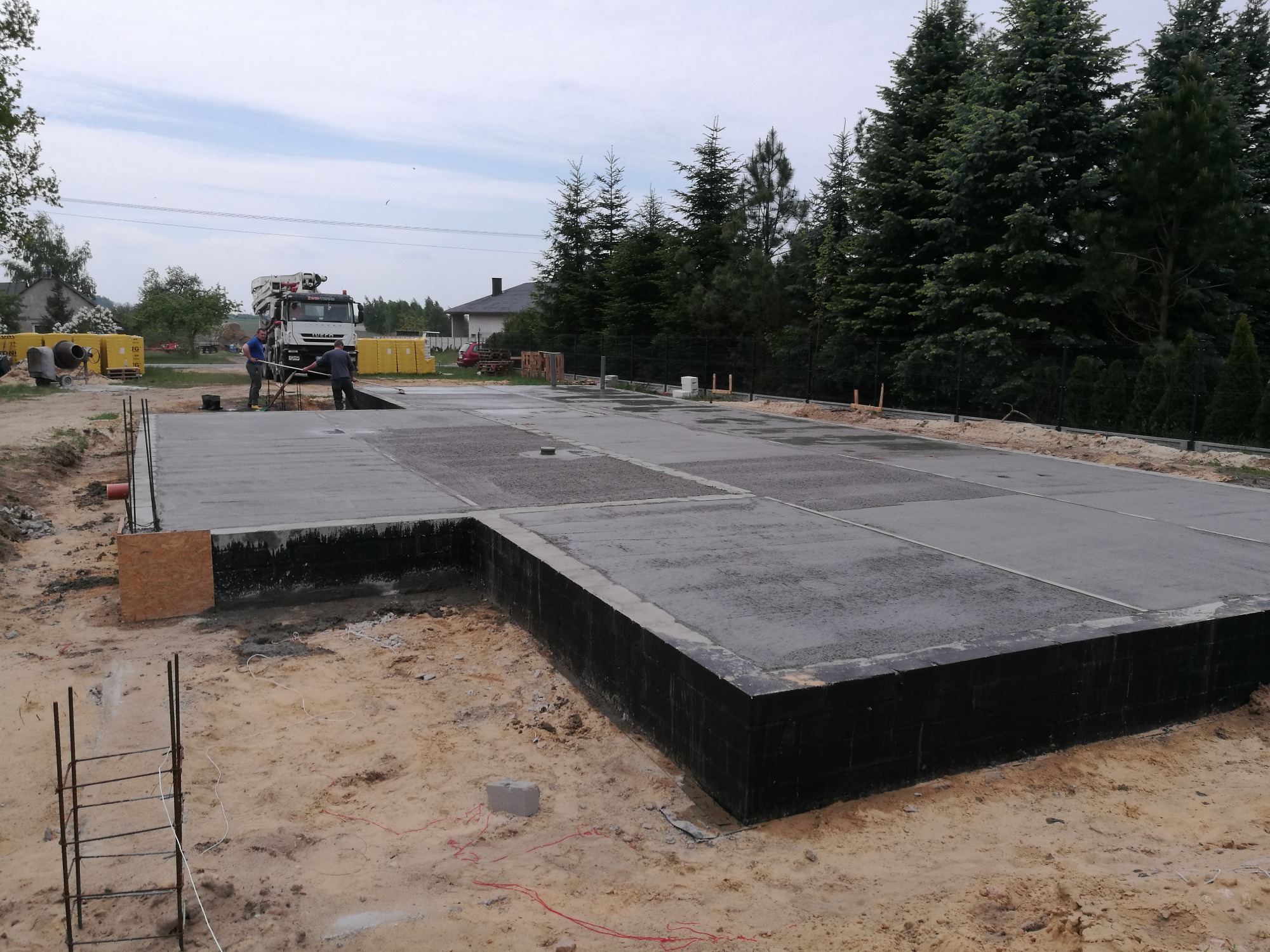 Wylany betonowy fundament pod budynek z widocznym szalunkiem i pracownikami przy betoniarce w tle.