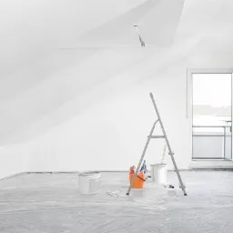 Pokój na poddaszu przygotowany do malowania: podłoga zabezpieczona folią, białe ściany, drabina, wiaderka z farbą i akcesoriami malarskimi.