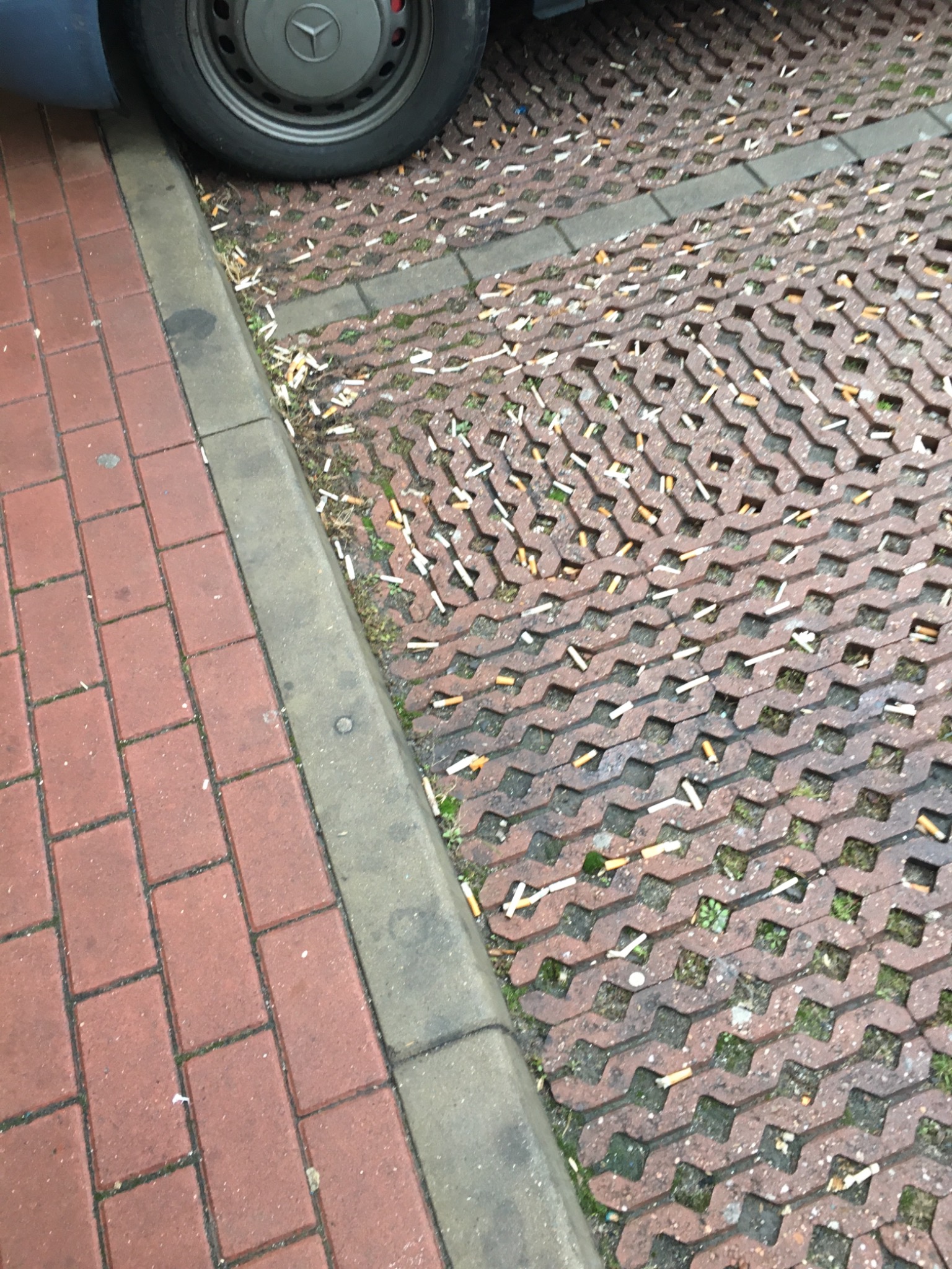 Zanieczyszczona kostka ażurowa i chodnik z czerwonej cegły przy samochodzie, zaśmiecone niedopałkami papierosów.