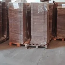 Stosy płaskich kartonów na paletach, owinięte folią stretch, składowane w magazynie z betonową podłogą i białymi ścianami.