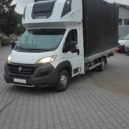 Biały samoch&oacute;d dostawczy marki Fiat z czarną plandeką na skrzyni ładunkowej, zaparkowany na szarym brukowanym placu. Widoczny napis 'LAMAR' na nadbud&oacute;wce kabiny.