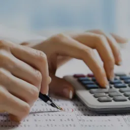 Osoba w białej koszuli używa kalkulatora i długopisu z piórem na dokumencie z danymi liczbowymi, zakreślając wynik na raporcie.