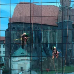 Dwóch alpinistów z żółtymi kaskami i sprzętem zabezpieczającym, zawieszonych na linach, czyści szklaną elewację budynku, w której odbija się zabytkowy kościół z czerwonym dachem.