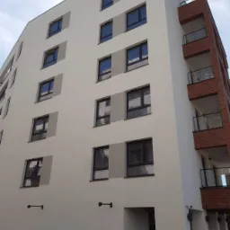 Wielopiętrowy budynek z ocieploną elewacją w odcieniach bieli i szarości, z fragmentem elewacji z czerwonej cegły i balkonami z przezroczystymi balustradami, widok z dołu na błękitne niebo.