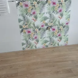 Ściana wyłożona płytkami z motywem tropikalnych kwiatów i liści, sąsiadująca z panelami podłogowymi imitującymi drewno, oraz fragment białego grzejnika drabinkowego.