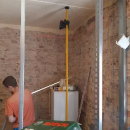 Wnętrze w trakcie prac remontowych: widoczne metalowe profile konstrukcyjne, czerwona linia lasera wyznaczająca poziom, odsłonięta ceglana ściana i częściowo wykończona powierzchnia, pudełko...