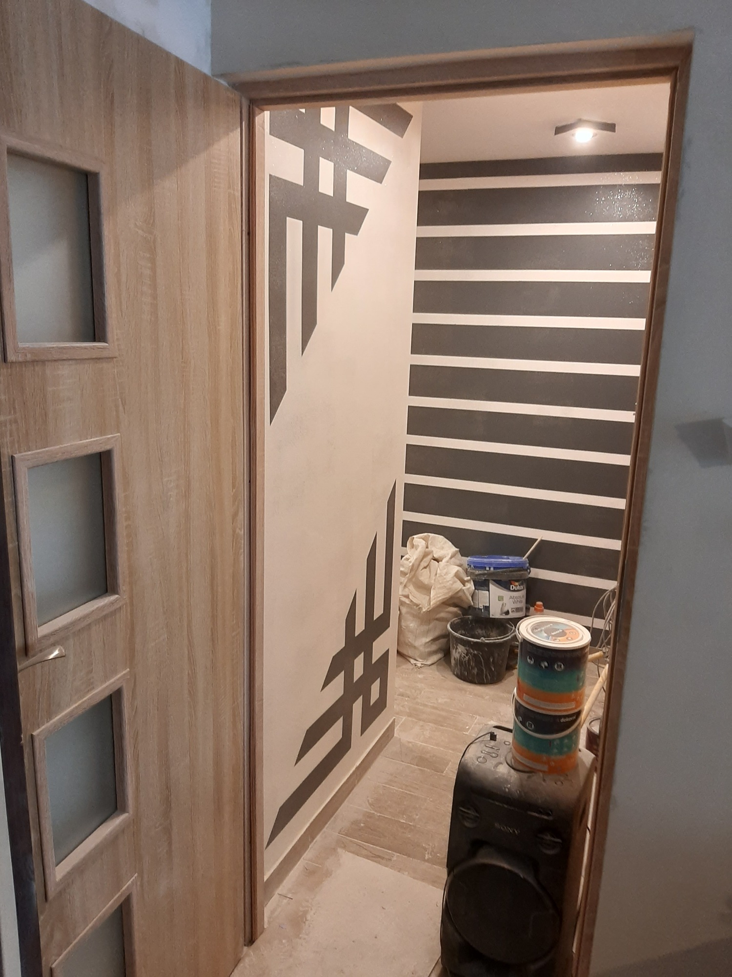 Wnętrze w trakcie malowania: drzwi z przeszkleniami, ściana z geometrycznym wzorem i poziome pasy, widoczne puszki z farbą i głośnik Sony na podłodze.