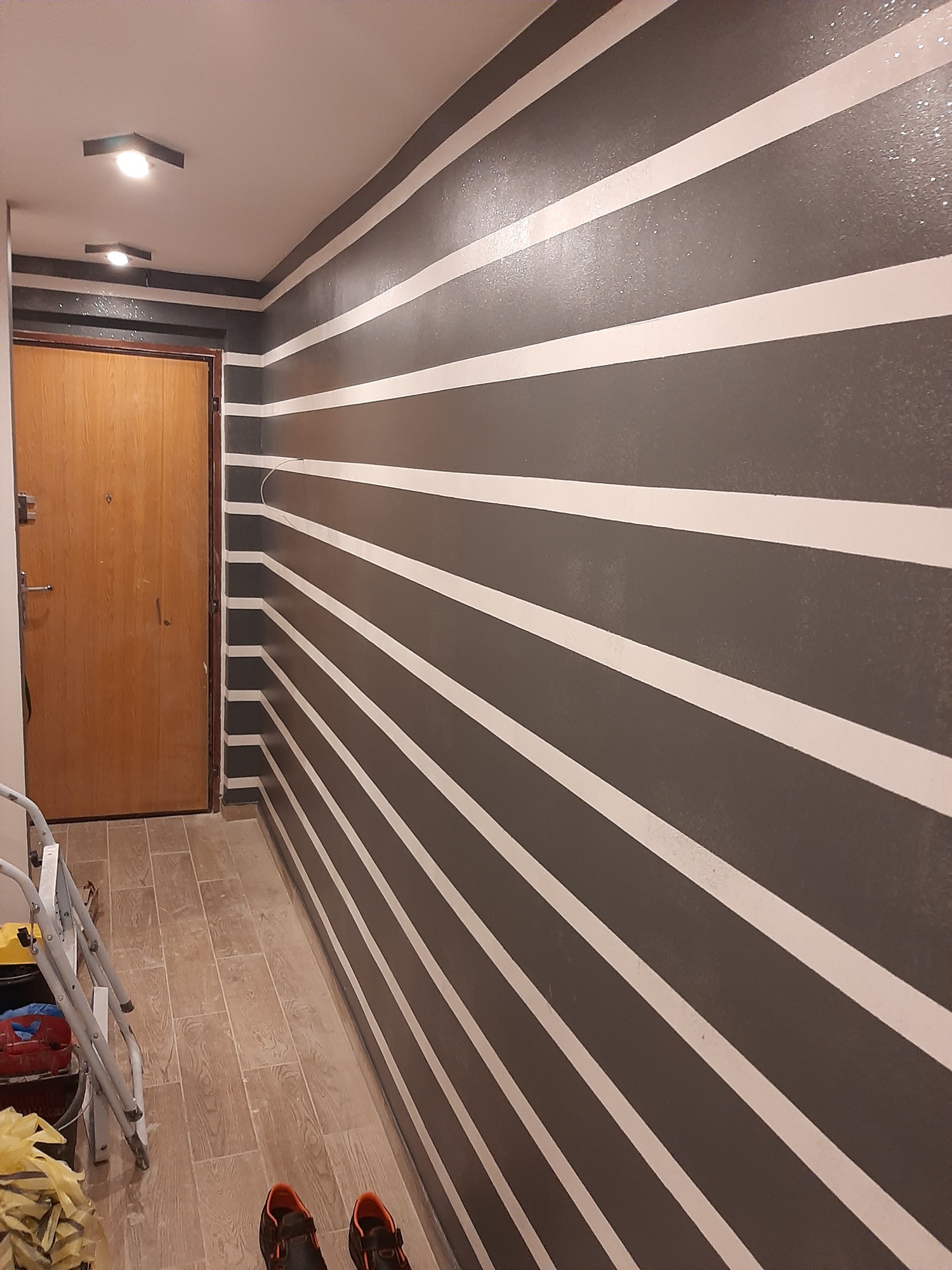 Korytarz po malowaniu ścian w poziome, szare i białe pasy, widoczna drewniana podłoga i część drabiny malarskiej.