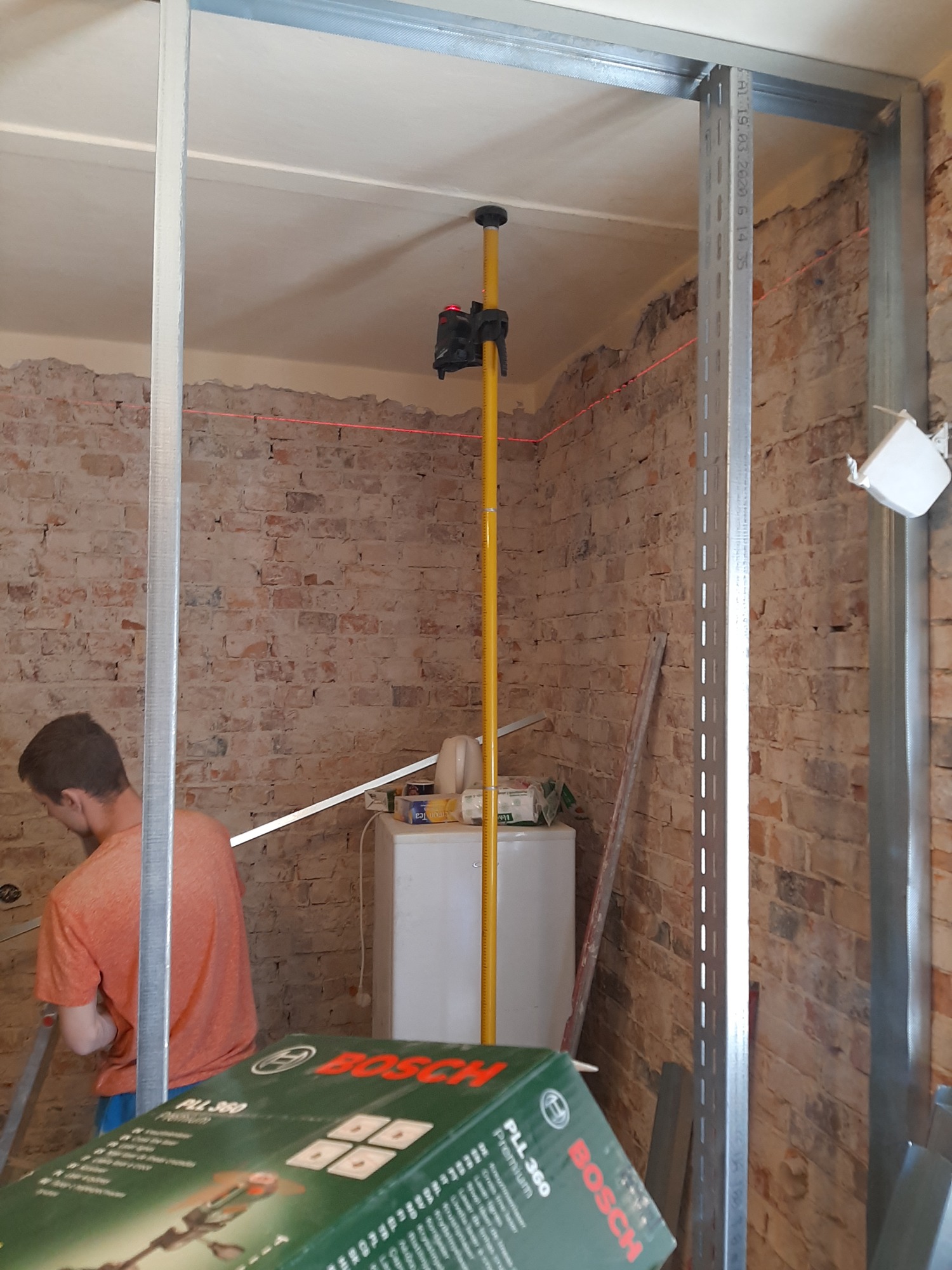 Wnętrze w trakcie prac remontowych: widoczne metalowe profile konstrukcyjne, czerwona linia lasera wyznaczająca poziom, odsłonięta ceglana ściana i częściowo wykończona powierzchnia, pudełko...