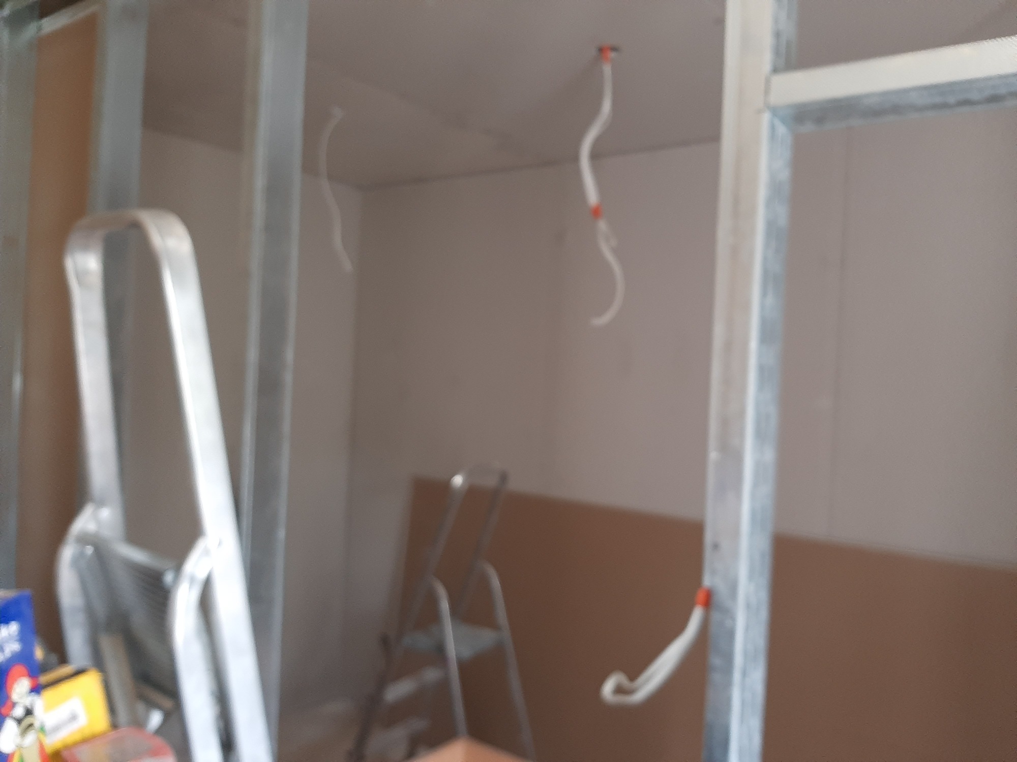 Wnętrze w trakcie prac wykończeniowych: widoczne profile metalowe konstrukcji ścian działowych, przewody elektryczne wystające z sufitu i ściany, oraz drabina aluminiowa w tle.