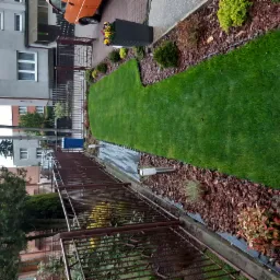 Widok na świeżo założony trawnik z rolki, obłożony korą, z widocznym ogrodzeniem i fragmentem budynku w tle, całość utrzymana w stonowanej kolorystyce.