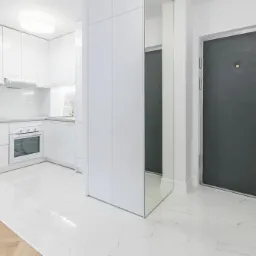 Nowoczesne, białe wnętrze mieszkania: kuchnia z marmurowym blatem, szafki z złotymi uchwytami, szara tapicerowana krzesło, ciemnoszare drzwi wejściowe z złotą klamką i podłoga z białego marmuru...