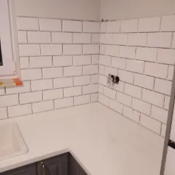 Białe, prostokątne płytki ułożone na ścianie nad blatem kuchennym, widoczne gniazdka elektryczne bez osprzętu, szare szafki pod blatem z złotymi uchwytami.