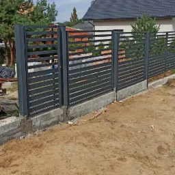 Nowoczesne, poziome, szare przęsła ogrodzeniowe, częściowo zamontowane na betonowej podmurówce, na tle domu z ciemnym dachem i zieleni