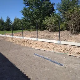 Wykop pod fundament ogrodzenia betonowego z zamontowanymi słupkami i widocznymi elementami szalunku oraz folią ochronną na profilu.