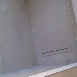 Nowoczesna kabina prysznicowa z odpływem liniowym, wykończona jasnymi, marmurkowymi płytkami ceramicznymi, widoczna szklana ściana z metalowymi okuciami.