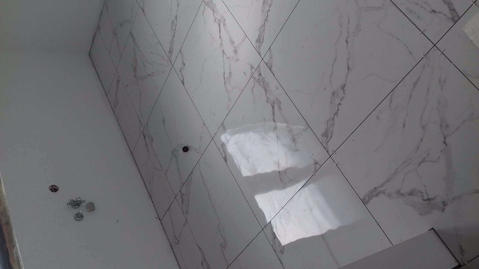 Błyszczące, białe płytki imitujące marmur, ułożone ukośnie na podłodze, widoczne krzyżyki dystansowe w fugach, fragment ściany z wystającymi zaworami.