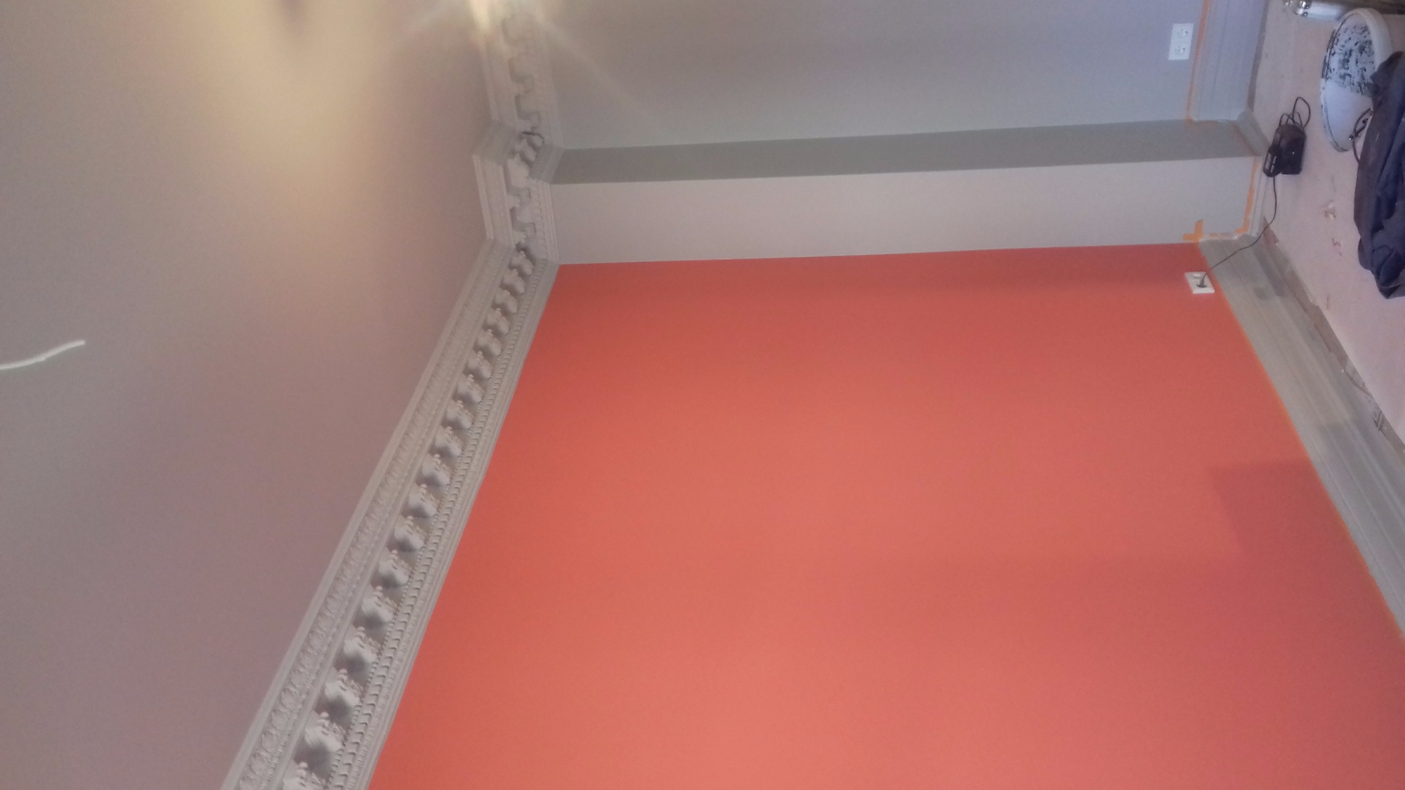 Ściana pomalowana na odcień koralowy, z dekoracyjnym gzymsem sufitowym i widocznym gniazdkiem elektrycznym, w trakcie prac wykończeniowych, widoczne zabezpieczenia podłogi.